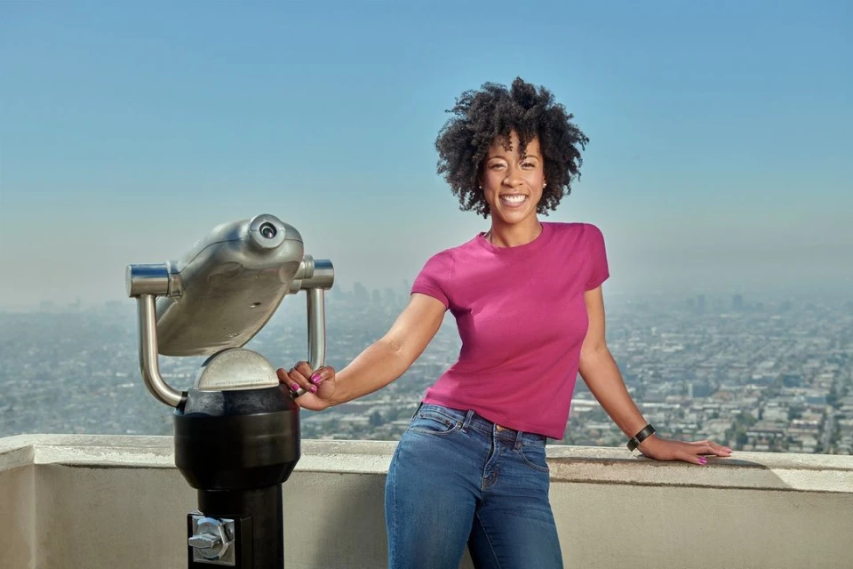 Smiling woman poses on rooftop near coin-operated binoculars, overlooking a sprawling cityscape under a clear blue sky during daytime.
